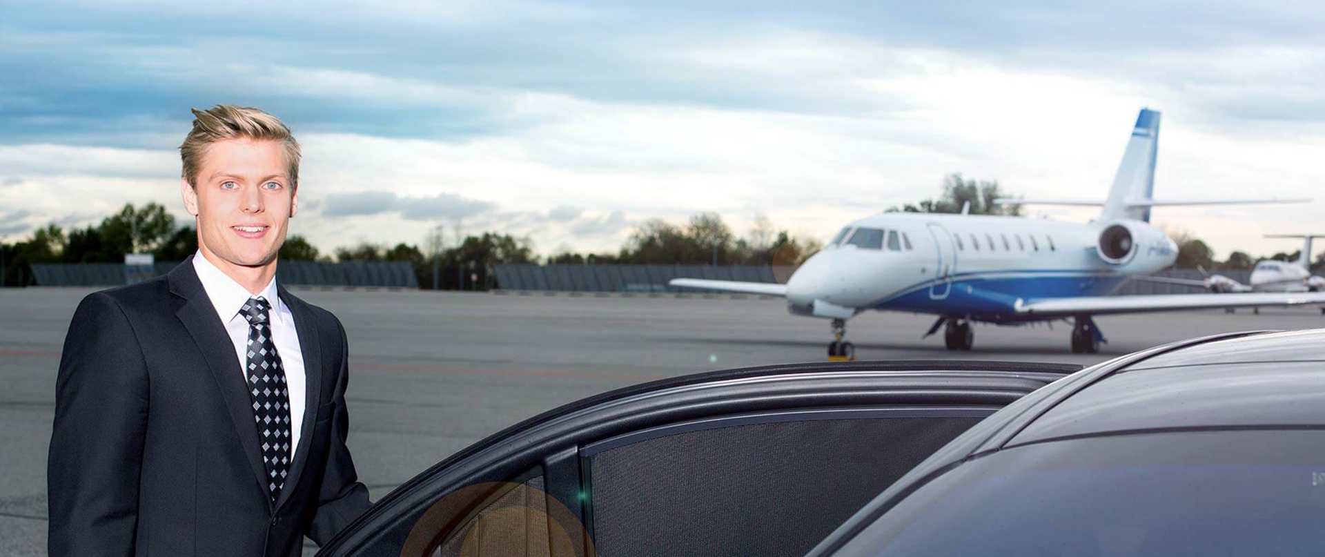 Ein Chauffeur öffnet die Tür einer Limousine, auf dem Rollfeld vor einem Business-Jet.